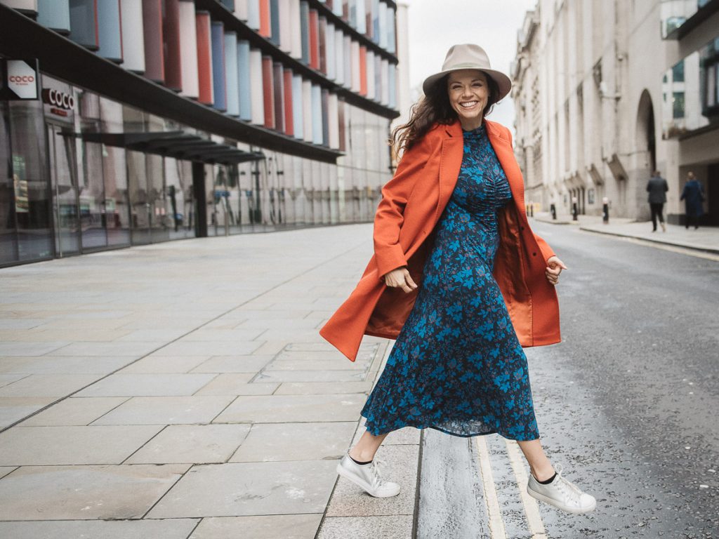 JustJerriJ: Jerri Jarmeh, photographer, smiling at camera in blue floral dress and orange jacket while walking on streets in downtown London, United Kingdom.